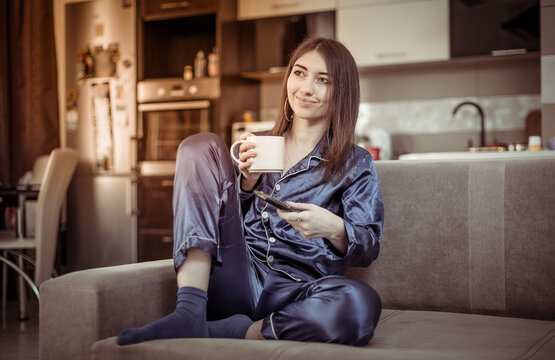 Young woman in nightie drinks coffee and watches TV while sitting on the sofa in the living room