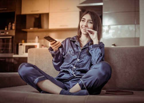 Young cheerful woman in a nightie switches TV channels with the remote control and eats, watching TV while sitting on the sofa in the living room