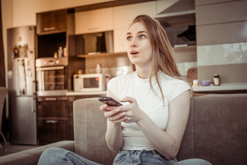 Keen woman looks at a TV screen and switches TV channels with a remote control, sitting on a sofa in a living room.
