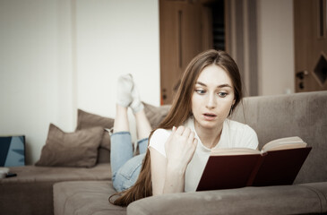 Young attractive woman lying on the sofa and reading interesting book in living room