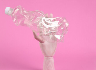 Wooden hand holds crumpled plastic bottle on pink background