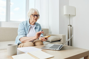 a beautiful elderly woman who is passionate about work is sitting at home in a bright apartment on a cozy sofa and checking finances looking at a laptop doing calculations on a calculator in her phone