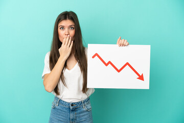 Young caucasian woman isolated on blue background holding a sign with a decreasing statistics arrow symbol with surprised expression