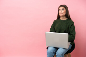 Young caucasian woman sitting on a chair with her laptop isolated on pink background and looking up