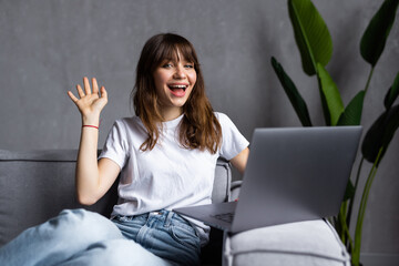 Happy young woman with toothy smile waving hand, looking at camera, sitting on couch at home,