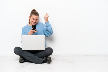 Young woman with a laptop sitting on the floor with phone in victory position