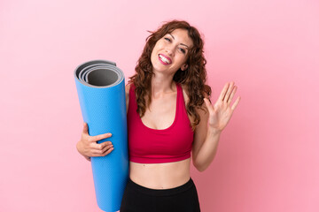 Young sport woman going to yoga classes while holding a mat isolated on pink background saluting with hand with happy expression