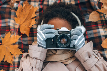 Cute African-American girl in fur headphones takes pictures with a camera lying on a blanket in an...