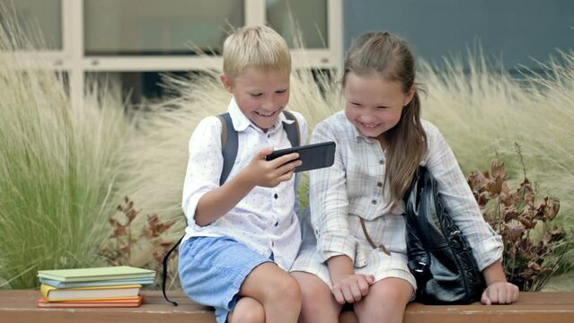 Two friends, a boy and a girl aged 8-9, are sitting on a bench in the schoolyard. Children look at the smartphone screen with interest.