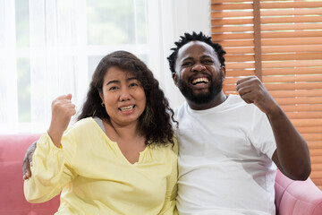 Obraz premium Happy diverse family African American male and asian female playing and watching tv while sitting on sofa in living room at home