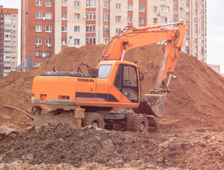 An orange excavator on a construction site against the background of a multi-storey building digs a trench. The excavator is in the process of operation.
