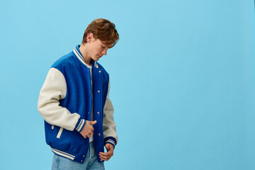 a handsome, attractive, young man student in a trendy blue bomber jacket stands with his head down and straightens the edge of his jacket on a light blue background. Horizontal photo with empty space