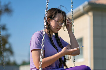 a teenager girl swings on a swing with a phone in her hands, communicates in social networks or learns online