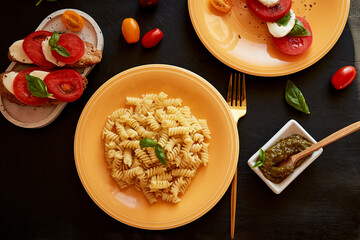 Fusilli pasta, caprese salad with tomato, basil, mozzarella appetizer with pesto on black rustic background. View from above. Italian dinner.