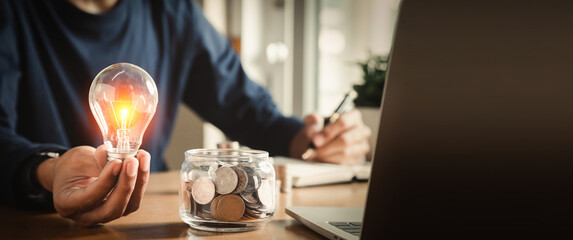 Businessman holding and putting lightbulb on coins stack on table for saving energy and saving money concept.