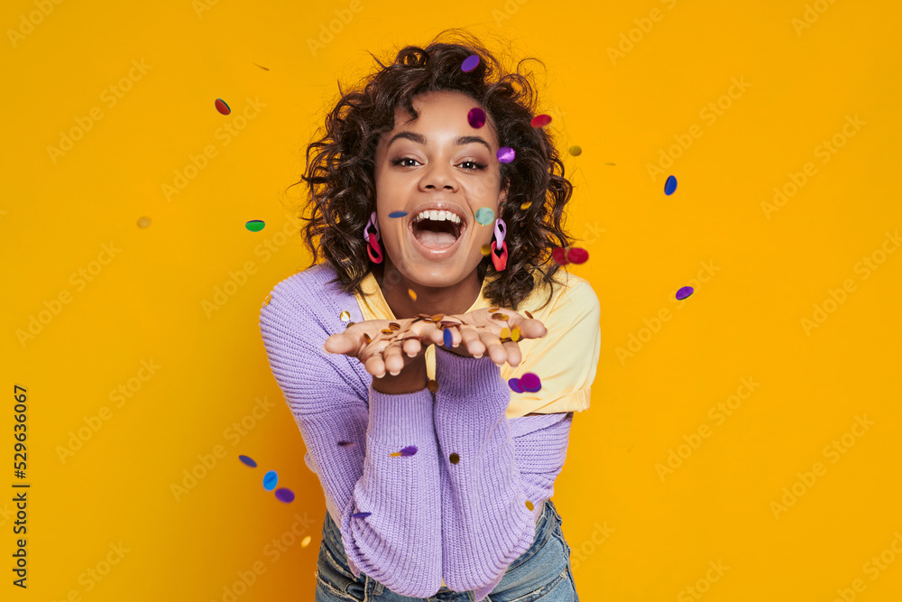 Wall mural Playful African woman blowing confetti and smiling while standing against yellow background