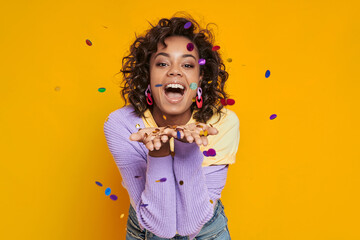 Playful African woman blowing confetti and smiling while standing against yellow background