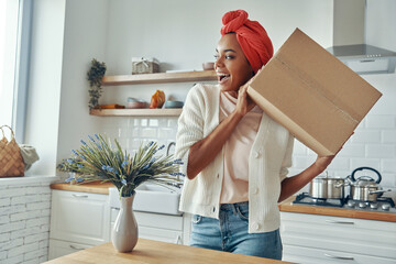 Excited African woman holding cardboard box near face and smiling while standing at kitchen