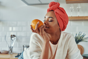 Attractive African woman in traditional headwear smelling oranges while standing at the kitchen - obrazy, fototapety, plakaty