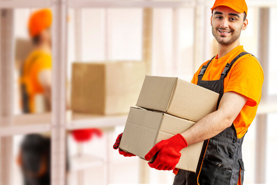 Young man holding cardboard package working in warehouse among racks and shelves. Delivery man with box. Staff laborer, orange uniform cap, t-shirt, coveralls service moving delivering orders goods