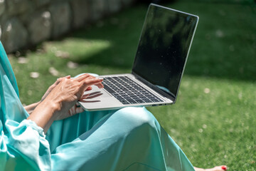 a young beautiful woman with blond curly hair in glasses and a blue dress sits on the grass in nature and uses a laptop,