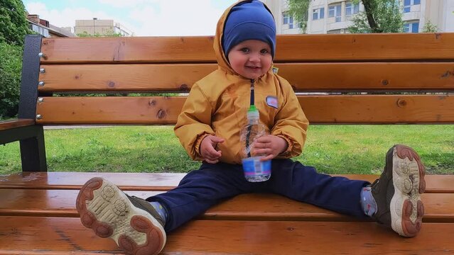 A cheerful child sits and moves his legs on a park bench. Boy 2 years old holding a bottle of water and laughing. Copy space - vacation concept, family walk, thirst, good mood, spring weather