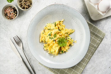 Breakfast scrambled eggs with green herbs, parsley in deep grey bowl on grey neutral table, vintage fork and knife, top view