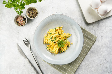 Breakfast scrambled eggs with green herbs, parsley in deep grey bowl on grey neutral table, vintage fork and knife, top view - obrazy, fototapety, plakaty