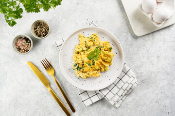 breakfast scrambled eggs with green herbs, parsley in white plate on grey neutral table, golden fork and knife, top view - obrazy, fototapety, plakaty