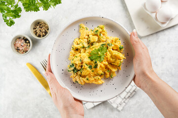 Female hands holding breakfast scrambled eggs with green herbs, parsley in white plate on grey...