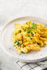 Breakfast scrambled eggs with green herbs, parsley in white plate on grey neutral table, golden fork and knife