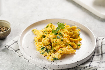 Breakfast scrambled eggs with green herbs, parsley in white plate on grey neutral table, golden fork and knife