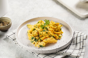 Breakfast scrambled eggs with green herbs, parsley in white plate on grey neutral table, golden fork and knife