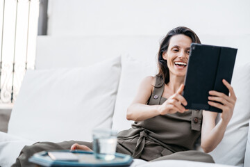 A young brunette latina woman sitting on a sofa smiles while video conferencing with her tablet.