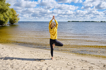 Fototapeta na wymiar A beautiful young yoga instructor does vrikshasana, stands on one leg and raises her folded palms up. Yoga outdoors by the sea or ocean. Healthy active lifestyle. Health and sport concept 