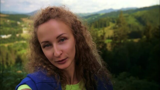 Portrait of Young Woman take a selfie on the mountains background.Curly Female blogger doing video call using smartphone against nature landscape.
