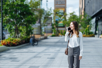 Smiling Asian curly business woman wearing trendy walks down the central city street and uses her phone. Pretty summer woman in white shirt walks down the street looking at her mobile phone