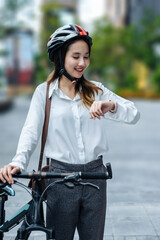 Naklejka premium Young Asian businesswoman wearing helmet and standing on a city street with bicycle looking at the watch.