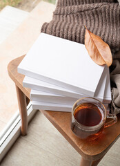 Stack of white blank books with autumn leaves and cup of hot tea on old wooden chair, mockup design