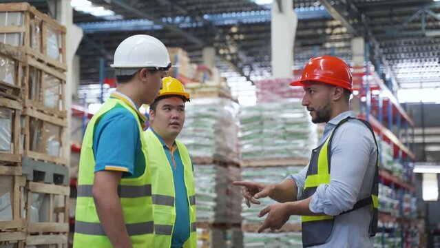 4K, Large warehouse manager is advising two employees standing in warehouse on occupational safety to wear reflective vests and protective hard hats and train workers in morning. Order subordinates