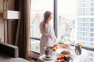 Woman in bathrobe eating breakfast near window with city view