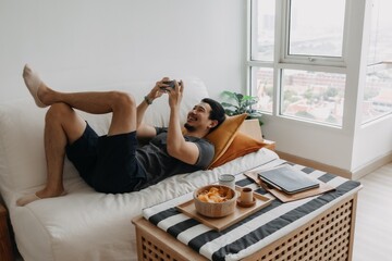 Lazy happy asian man playing mobile game while he should have work from home. - Powered by Adobe