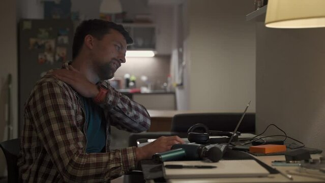 Neck pain, cervical osteochondrosis. Male student studying until night at computer with neck pain. Tired guy kneads neck with pistol shock massager while working on laptop. Muscle recovery and massage