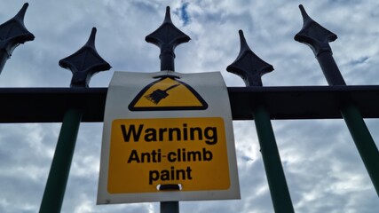 A low angle landscape shot of a yellow warning sign reading Warning anti climb paint attached to a fence with sharp tips against a cloudy sky.