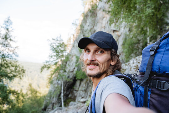 Curly guy in a cap takes a selfie on his phone, a hiker in the mountains, a backpack on his back, a bearded hipster on a hike, a black hat, a hat