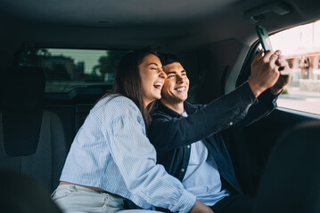 Happy couple on road trip. Young man and woman sitting on rear seat of car looking at phone smiling on a summer day.