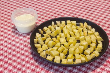 A traditional dish of potatoes with aioli in a plastic bowl is served on a table with a checkered tablecloth. Holidays and tapas.