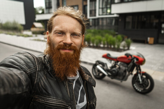 Redheaded, bearded biker with no helmet photographing himself in front of a motorcycle