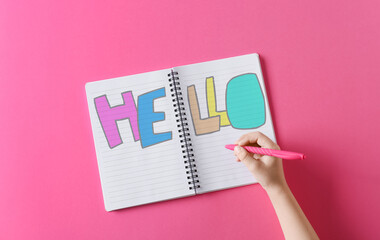 Child writing word HELLO in notebook on pink background
