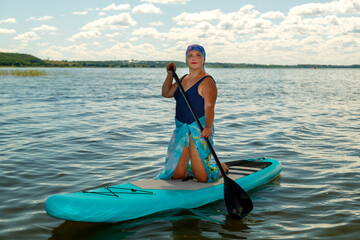 A Jewish woman in a headscarf in a pareo on her knees on a SUP board swims in the lake on a sunny day.